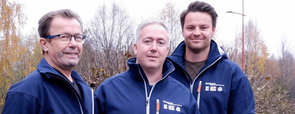 Teamet i Øystein Opås Takstforretning står utendørs i firmajakker og smiler mot kamera, med høstfargede trær i bakgrunnen.